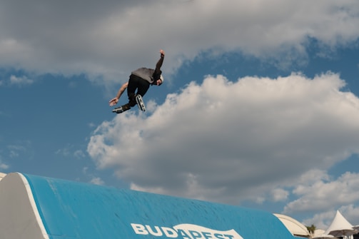 A person wearing inline skates performs an acrobatic jump against a backdrop of a partly cloudy blue sky. The lower part of the image shows a blue ramp with the word 'BUDAPEST' on it.