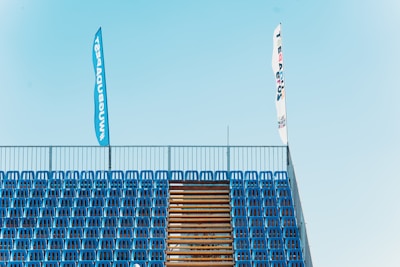 Rows of blue stadium seats are neatly arranged, separated by a central staircase. Two tall flags are visible above the seating area, one blue and one white.