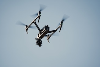 A black drone with four propellers is flying against a clear blue sky. The drone has a camera mounted underneath, and its rotors are clearly visible due to the motion blur.