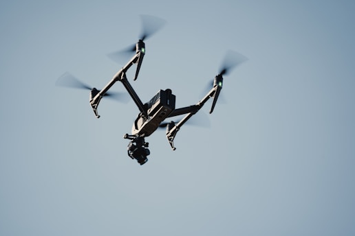 A black drone with four propellers is flying against a clear blue sky. The drone has a camera mounted underneath, and its rotors are clearly visible due to the motion blur.