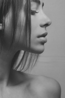 A monochrome close-up portrait of a person in profile, emphasizing their facial features. The image captures the smooth texture of the skin and the soft lines of the face, with hair gently cascading down. The person's eyes are closed, conveying a sense of calm and introspection.