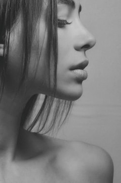 A monochrome close-up portrait of a person in profile, emphasizing their facial features. The image captures the smooth texture of the skin and the soft lines of the face, with hair gently cascading down. The person's eyes are closed, conveying a sense of calm and introspection.