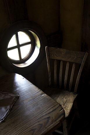 There is a rustic wooden chair next to a small wooden table. The table has a cloth partially on it, and both pieces of furniture have a distinct natural wood grain. A round window with a cross design is seen on the left side, allowing some light into the otherwise dark room.