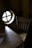 There is a rustic wooden chair next to a small wooden table. The table has a cloth partially on it, and both pieces of furniture have a distinct natural wood grain. A round window with a cross design is seen on the left side, allowing some light into the otherwise dark room.