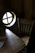 There is a rustic wooden chair next to a small wooden table. The table has a cloth partially on it, and both pieces of furniture have a distinct natural wood grain. A round window with a cross design is seen on the left side, allowing some light into the otherwise dark room.
