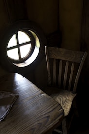There is a rustic wooden chair next to a small wooden table. The table has a cloth partially on it, and both pieces of furniture have a distinct natural wood grain. A round window with a cross design is seen on the left side, allowing some light into the otherwise dark room.
