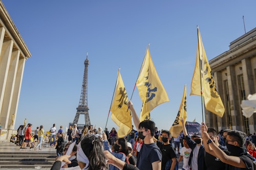 A group of people gathers near a famous landmark, holding yellow flags with black symbols. The scene is bustling with individuals, some wearing face masks, and the Eiffel Tower is visible in the background against a clear blue sky. The architecture of the adjacent building suggests a neoclassical style with tall columns.