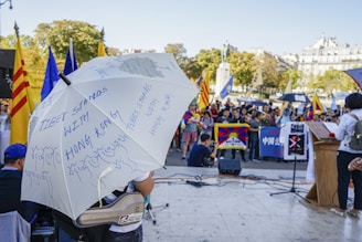 A public demonstration with a large group of people gathered outdoors. A prominent white umbrella is seen in the foreground, with messages written on it in blue ink, including 'Tibet Stands with Hong Kong.' The crowd holds various flags, including the Tibetan flag, and banners. A speaker addresses the crowd from a podium on the right side.
