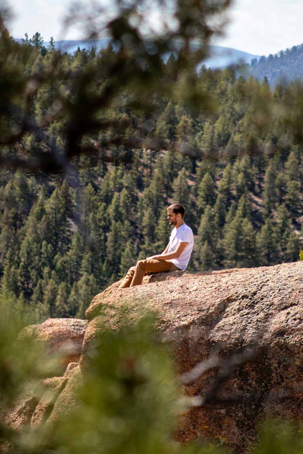 man wearing white crew-neck shirt and blue pants sitting on rock looking at mountain scenery