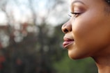 A close-up side profile of a woman with smooth skin and well-defined facial features. Her eyes are closed, and she has a calm expression. The background is blurred with natural colors, creating a soft and serene atmosphere.