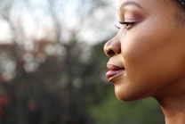 A close-up side profile of a woman with smooth skin and well-defined facial features. Her eyes are closed, and she has a calm expression. The background is blurred with natural colors, creating a soft and serene atmosphere.