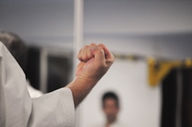 A close-up of a clenched fist, with part of a person's arm visible. The background is blurred, highlighting the focus on the gesture of the fist, which is likely in a martial arts setting given the context of attire.