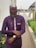 A person is wearing a traditional outfit with a purple color scheme, featuring a patterned cap and matching shirt with intricate designs along the chest. They are adjusting a watch on their wrist, standing in an outdoor setting with a walkway lined by green shrubs.