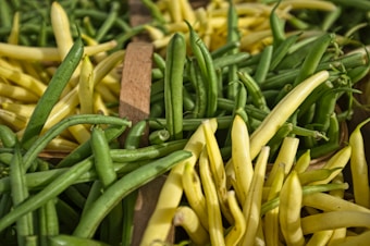 A close-up view of an assortment of fresh green and yellow beans, displayed in a market setting. The beans are piled together, showing their vibrant colors and natural textures. The lighting highlights their fresh and crisp appearance.