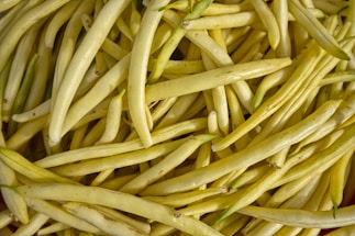 A close-up view of numerous yellow beans piled together, showcasing their slender and elongated shape. The beans have a smooth texture and slight variations in color with patches of green on some.