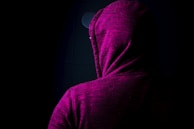 A person is wearing a dark magenta hoodie with the hood up, facing away from the camera. The background is dark, adding a sense of mystery to the scene. The fabric of the hoodie has a mottled texture.