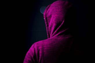 A person is wearing a dark magenta hoodie with the hood up, facing away from the camera. The background is dark, adding a sense of mystery to the scene. The fabric of the hoodie has a mottled texture.