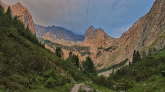 A scenic mountain landscape featuring rugged peaks and a lush green valley. Cable car lines run across the sky, connecting different points of the mountainous terrain. Pine trees are scattered along the slopes, adding to the natural beauty of the environment.