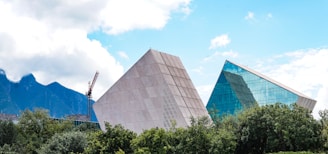 Two modern, angular buildings with a geometric design rise above lush green trees. One building has a smooth, gray facade, while the other features reflective blue glass panels. A crane is visible in the background, with mountains and a partly cloudy sky completing the backdrop.