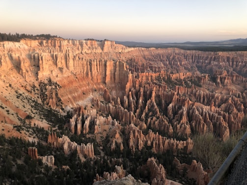 A breathtaking view of a vast canyon landscape characterized by orange and red-hued spires, cliffs, and hoodoos. The twilight sky adds a serene backdrop to the rugged terrain, while sparse vegetation is scattered among the rock formations.
