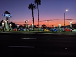 A car dealership is visible with multiple cars parked in rows under bright lights. The sky displays a gradient from orange to deep blue, indicating sunset or early evening. Palm trees are silhouetted against the colorful sky, and there are illuminated signs with the Ford logo.