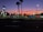 A car dealership is visible with multiple cars parked in rows under bright lights. The sky displays a gradient from orange to deep blue, indicating sunset or early evening. Palm trees are silhouetted against the colorful sky, and there are illuminated signs with the Ford logo.