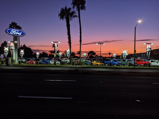 A car dealership is visible with multiple cars parked in rows under bright lights. The sky displays a gradient from orange to deep blue, indicating sunset or early evening. Palm trees are silhouetted against the colorful sky, and there are illuminated signs with the Ford logo.