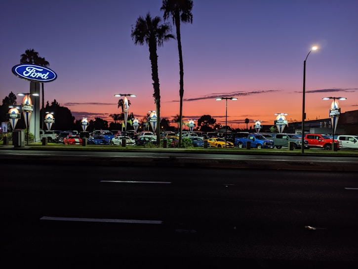 A car dealership is visible with multiple cars parked in rows under bright lights. The sky displays a gradient from orange to deep blue, indicating sunset or early evening. Palm trees are silhouetted against the colorful sky, and there are illuminated signs with the Ford logo.