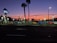 A car dealership is visible with multiple cars parked in rows under bright lights. The sky displays a gradient from orange to deep blue, indicating sunset or early evening. Palm trees are silhouetted against the colorful sky, and there are illuminated signs with the Ford logo.
