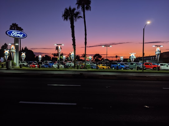 A car dealership is visible with multiple cars parked in rows under bright lights. The sky displays a gradient from orange to deep blue, indicating sunset or early evening. Palm trees are silhouetted against the colorful sky, and there are illuminated signs with the Ford logo.