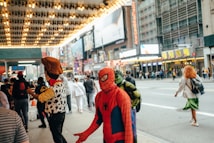 A busy urban street scene features people dressed in costumes of popular fictional characters. The background includes buildings with bright lights and advertisements typical of a city center. Pedestrians walk by as costumed figures interact.