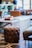 A cozy interior setting featuring a tufted leather ottoman with a dark, rich finish. The room has a mix of modern and rustic elements, including a wooden coffee table with books on top, patterned tiled flooring, and comfortable seating. Blurred figures are seen in the background, creating a relaxed, casual atmosphere.
