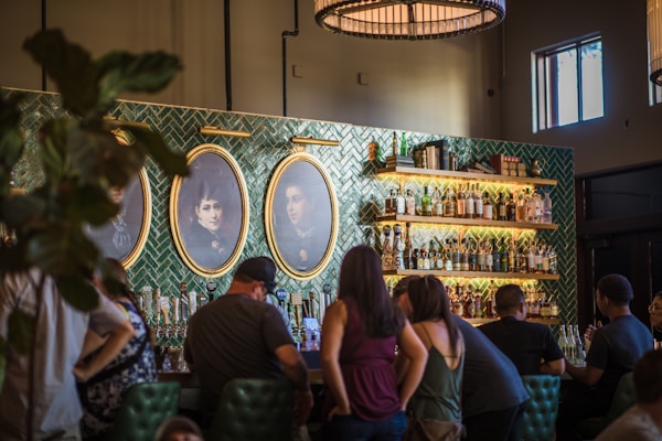 A cozy bar scene with a group of people gathered along a counter. The bar's backdrop features shelves filled with various bottles of liquor and books, set against an ornate green tiled wall. Above the shelves are two large antique-style portraits framed in gold. Soft lighting creates a warm and inviting atmosphere.