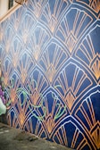 A wall mural features an intricate geometric pattern with fan-like shapes outlined in gold against a dark blue background. The design includes stylized green vine elements that add a touch of nature. The mural extends along a street, providing an eye-catching artistic presence.