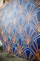A wall mural features an intricate geometric pattern with fan-like shapes outlined in gold against a dark blue background. The design includes stylized green vine elements that add a touch of nature. The mural extends along a street, providing an eye-catching artistic presence.