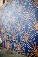 A wall mural features an intricate geometric pattern with fan-like shapes outlined in gold against a dark blue background. The design includes stylized green vine elements that add a touch of nature. The mural extends along a street, providing an eye-catching artistic presence.