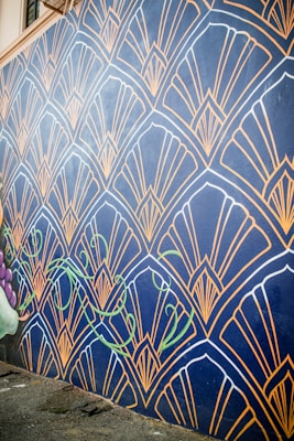 A wall mural features an intricate geometric pattern with fan-like shapes outlined in gold against a dark blue background. The design includes stylized green vine elements that add a touch of nature. The mural extends along a street, providing an eye-catching artistic presence.