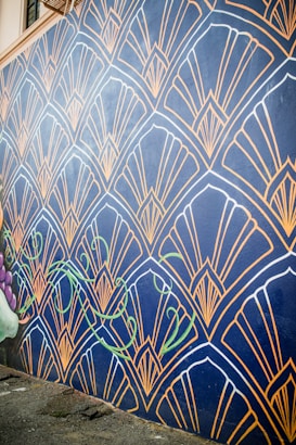 A wall mural features an intricate geometric pattern with fan-like shapes outlined in gold against a dark blue background. The design includes stylized green vine elements that add a touch of nature. The mural extends along a street, providing an eye-catching artistic presence.