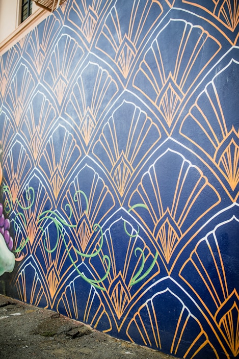 A wall mural features an intricate geometric pattern with fan-like shapes outlined in gold against a dark blue background. The design includes stylized green vine elements that add a touch of nature. The mural extends along a street, providing an eye-catching artistic presence.