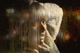 A woman appears contemplative, gazing out through a rain-streaked window. Her expression suggests introspection, while the dim lighting and reflections cast a melancholic ambiance. The raindrops create a textured, moody overlay.