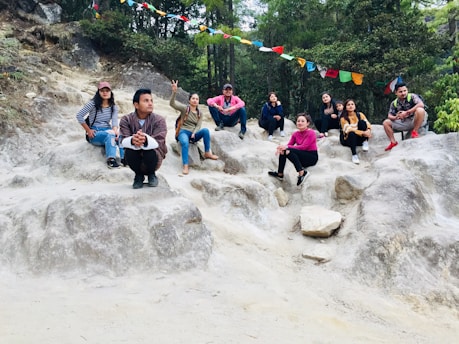 A group of people sitting on rocky terrain surrounded by greenery. They are casually dressed, and some are posing or making gestures. Colorful prayer flags are strung above them, adding a vibrant detail to the natural setting.