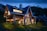 A modern wooden house with large windows and a gabled roof sits surrounded by well-maintained gardens under a twilight sky. Warm lights illuminate both the interior and exterior, creating a cozy and inviting atmosphere. The house has an elegant and rustic architectural style, with a stone chimney and landscaped pathway leading to the entrance.