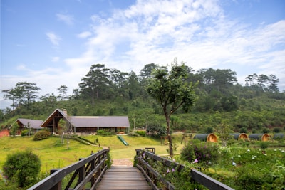 A rustic cabin with a sloping roof is situated amidst a lush green landscape, surrounded by trees and rolling hills. A wooden bridge leads the viewer's eye towards the cabin. In the vicinity, small, semi-circular glamping pods are visible, blending harmoniously with the natural surroundings. The sky is partly cloudy, adding a serene and tranquil atmosphere.