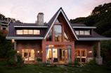 A modern house with triangular roofs and a large glass window displays warm indoor lighting. The exterior is made of wooden panels with a stone chimney. Surrounding the structure are lush green plants and a well-maintained garden. The sky has a soft pink hue, suggesting the time is either sunrise or sunset.