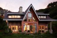 A modern house with triangular roofs and a large glass window displays warm indoor lighting. The exterior is made of wooden panels with a stone chimney. Surrounding the structure are lush green plants and a well-maintained garden. The sky has a soft pink hue, suggesting the time is either sunrise or sunset.