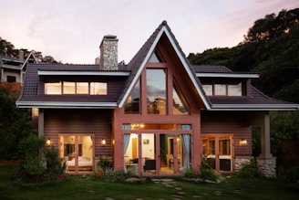 A modern house with triangular roofs and a large glass window displays warm indoor lighting. The exterior is made of wooden panels with a stone chimney. Surrounding the structure are lush green plants and a well-maintained garden. The sky has a soft pink hue, suggesting the time is either sunrise or sunset.