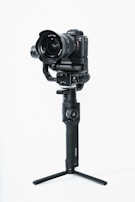 A professional camera mounted on a stabilizing gimbal with a tripod base. The camera has a large lens and brand markings can be seen. The gimbal is handheld with a grip designed for stability during video shooting.