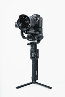 A professional camera mounted on a stabilizing gimbal with a tripod base. The camera has a large lens and brand markings can be seen. The gimbal is handheld with a grip designed for stability during video shooting.