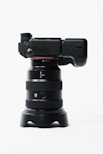 A professional digital camera with a large lens is positioned vertically. The camera body is black with a textured grip and various controls on the side. The lens has a wide barrel with multiple rings and a red 'G' logo. The setup is placed on a minimalistic white background, emphasizing the sleek design of the camera.