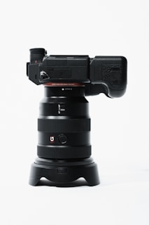 A professional digital camera with a large lens is positioned vertically. The camera body is black with a textured grip and various controls on the side. The lens has a wide barrel with multiple rings and a red 'G' logo. The setup is placed on a minimalistic white background, emphasizing the sleek design of the camera.